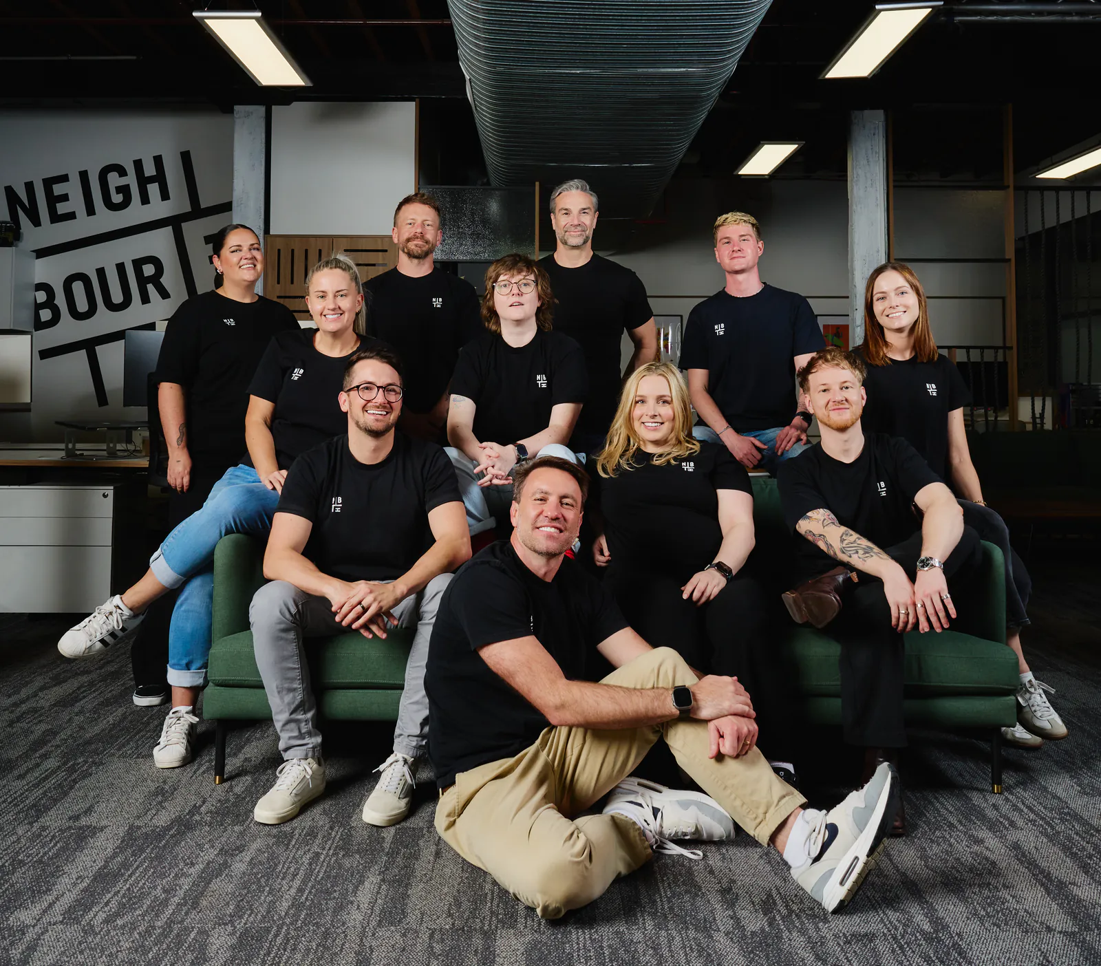 Eleven of the Neighbourhood crew on and around the green sofa in the studio, under the exposed ceiling
