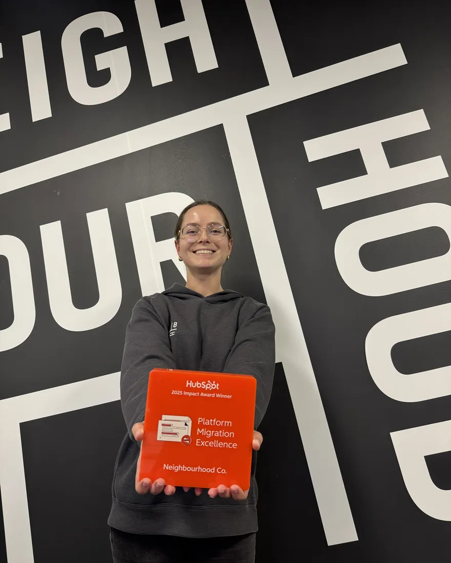 A Neighbour holding the 2025 HubSpot Impact Award for Platform Migration Excellence in front of the Neighbourhood wall