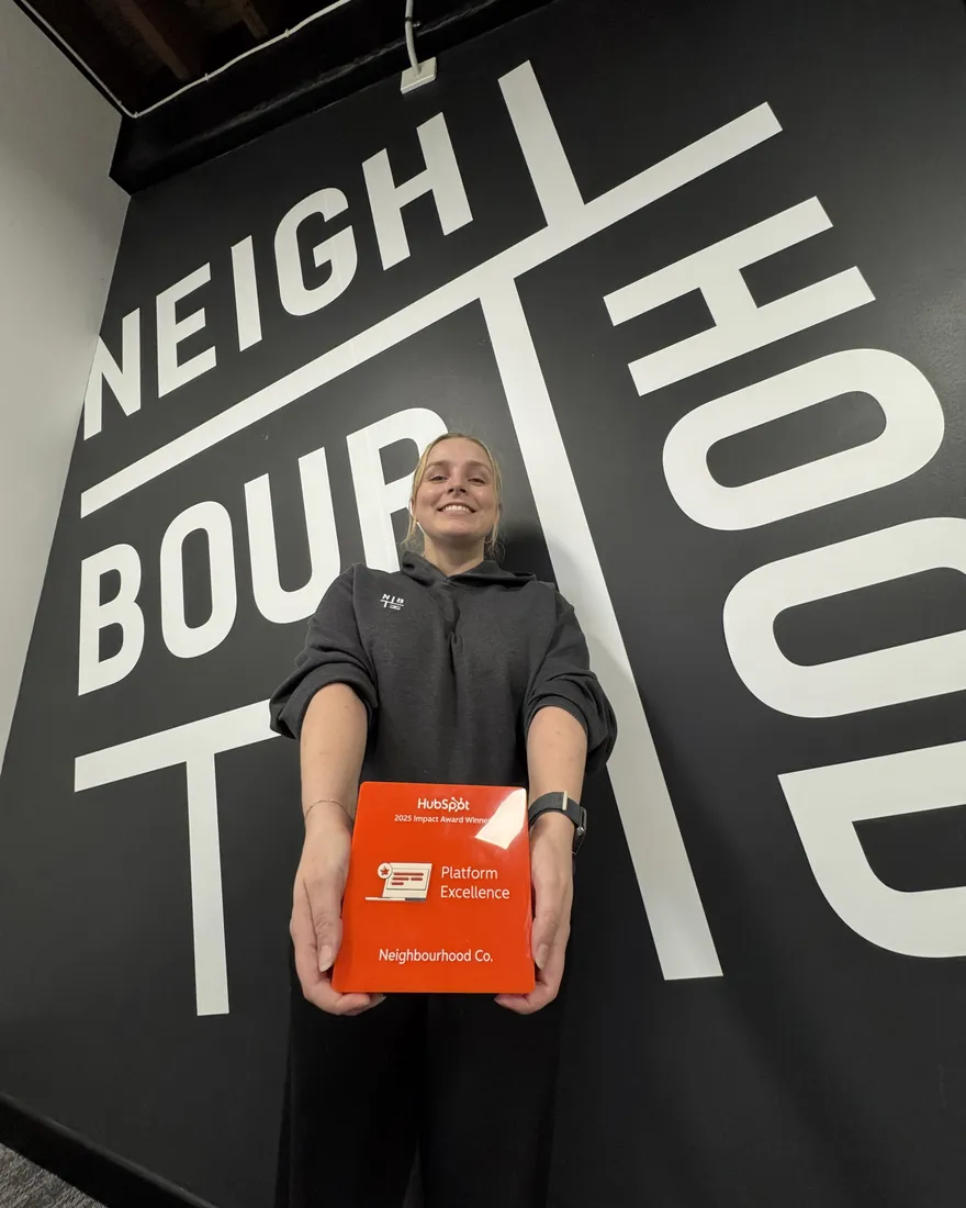 A Neighbour holding the 2025 HubSpot Impact Award for Platform Excellence in front of the Neighbourhood wall