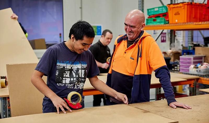 Two men working together at a packing bench in a warehouse, one taping a box, the other in a high visibility top