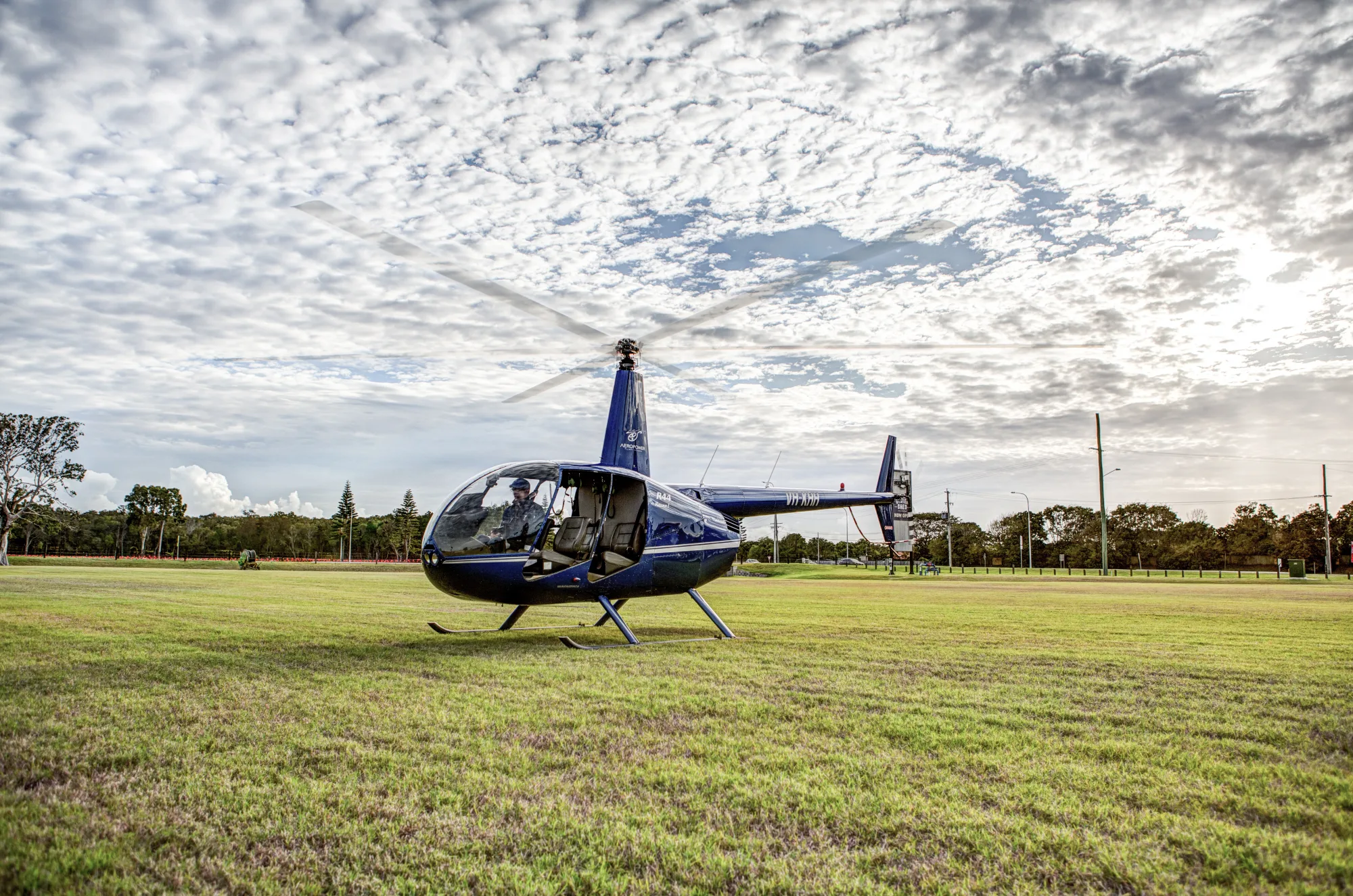 A blue Robinson R44 helicopter on a grass field under a cloudy sky