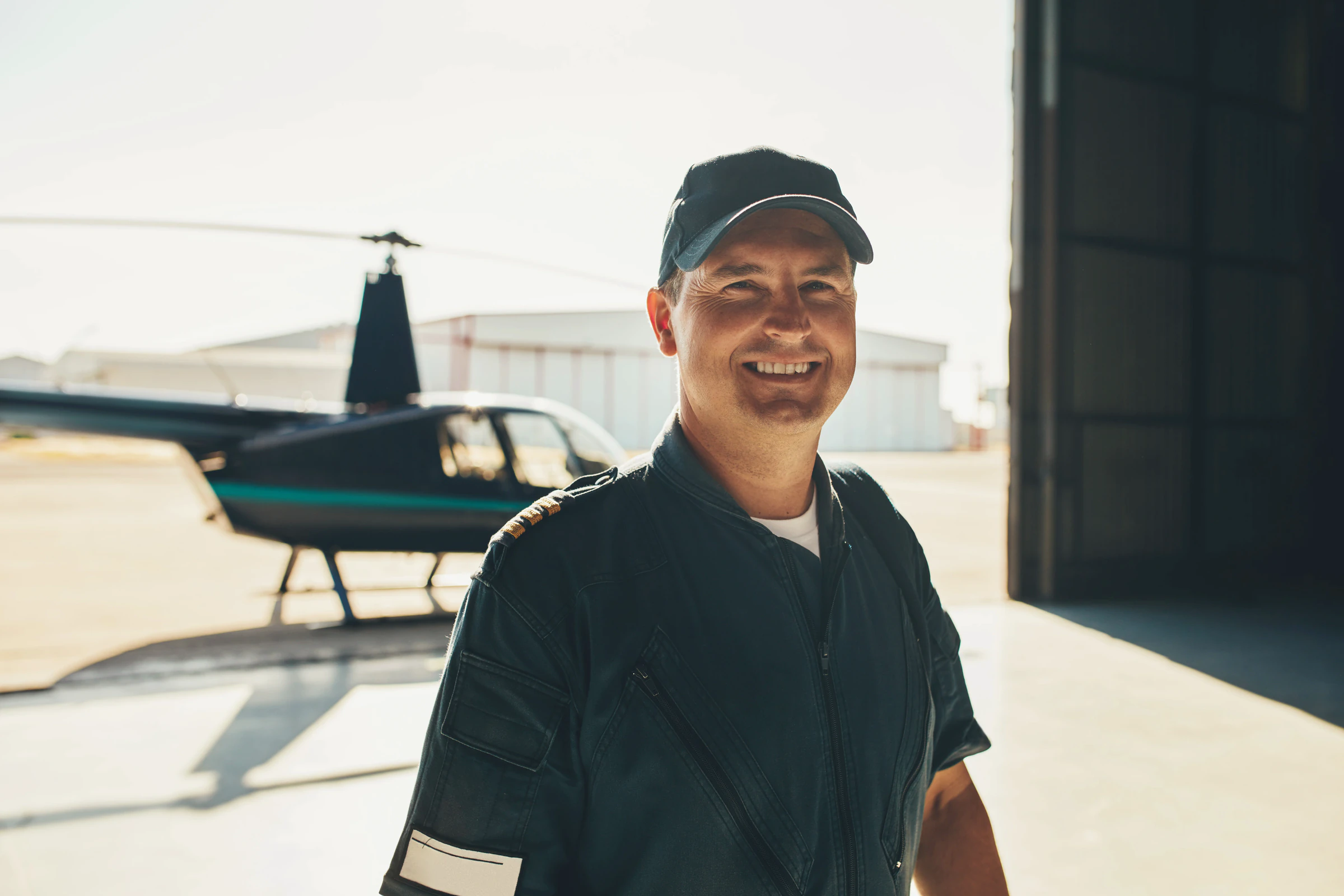 A smiling pilot in a hangar, with a helicopter behind him