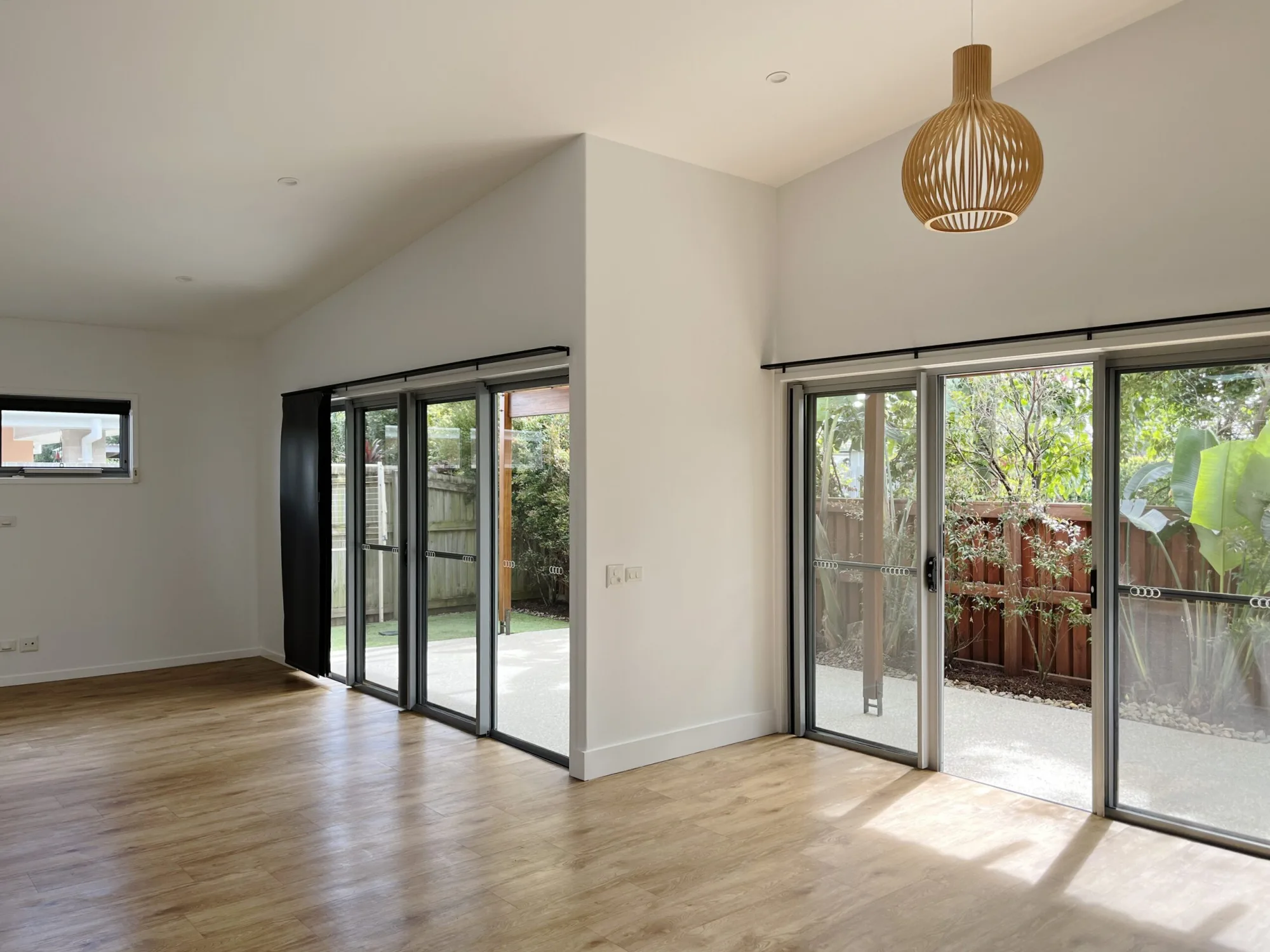 An empty lounge with a timber floor, a raked white ceiling, a woven pendant light and sliding doors to a garden