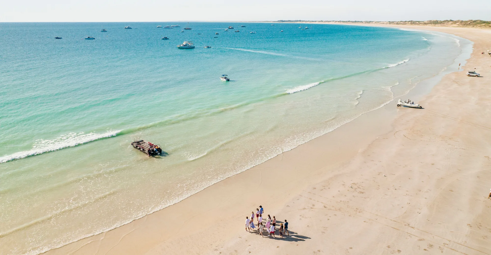 An aerial view of a long sandy beach and turquoise water, with boats moored offshore and a small group walking on the sand