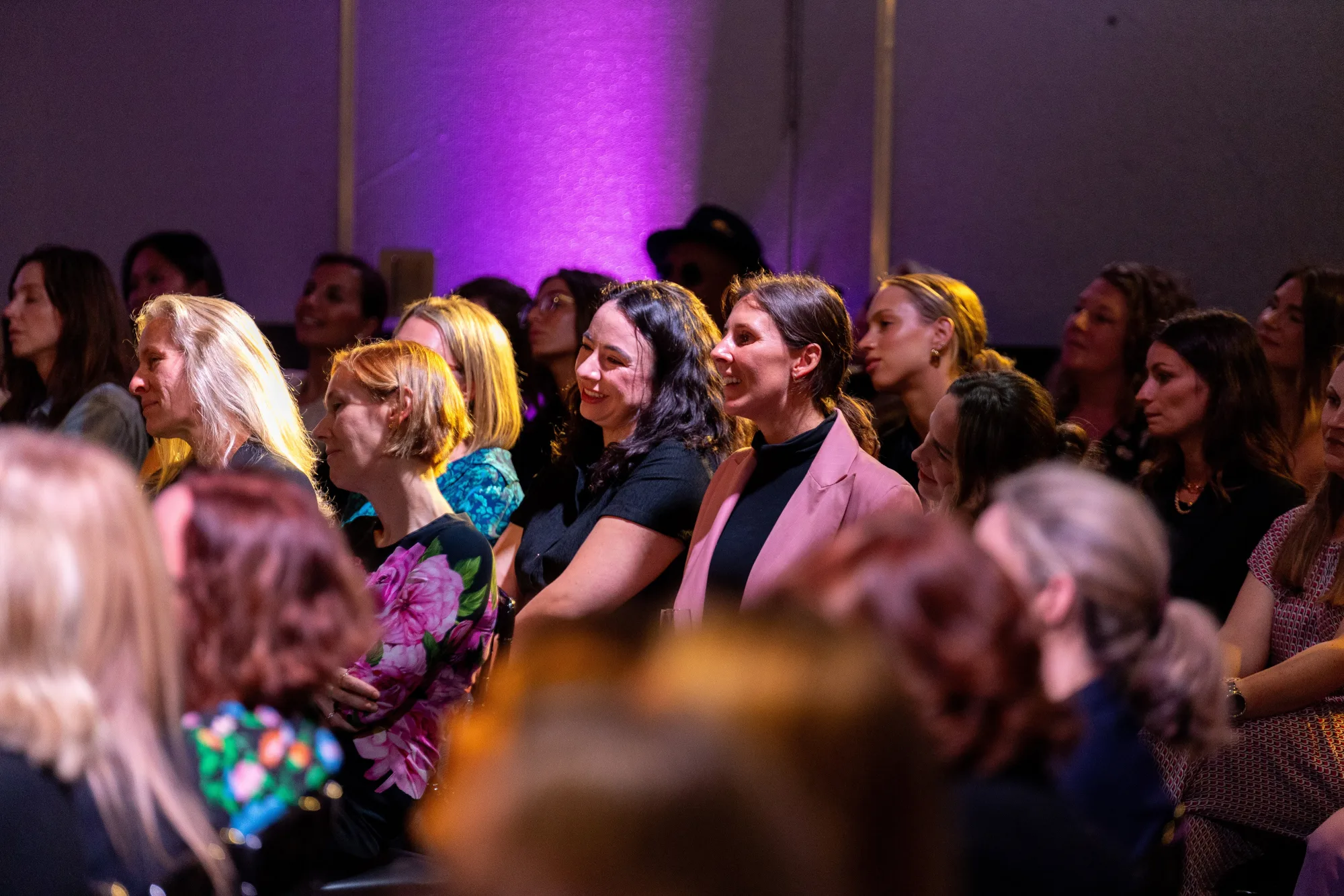 An audience of women seated at a CEW event under purple light, several smiling towards the stage