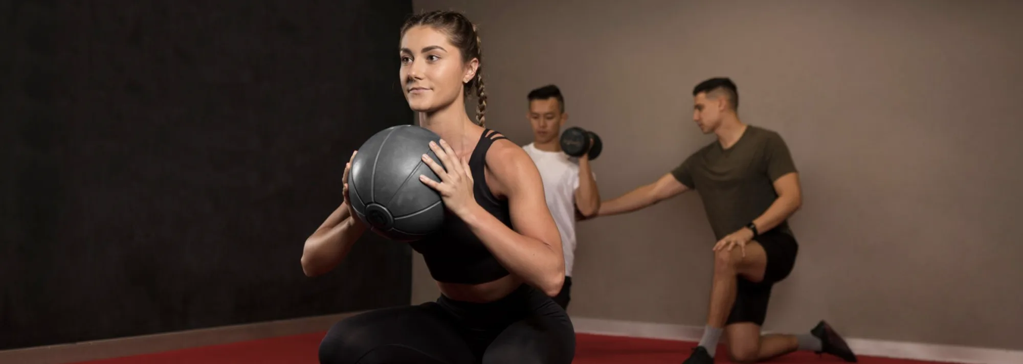 A woman holding a medicine ball in a squat, with a trainer coaching a man through a dumbbell curl behind her