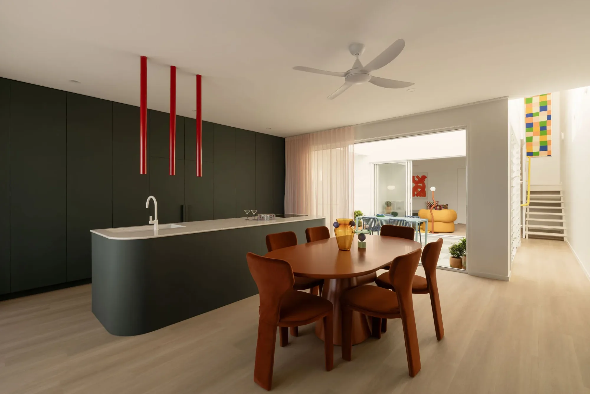 Kitchen and dining room in an AUSMAR display home: a dark curved island under three red pendants, a timber table and the courtyard beyond