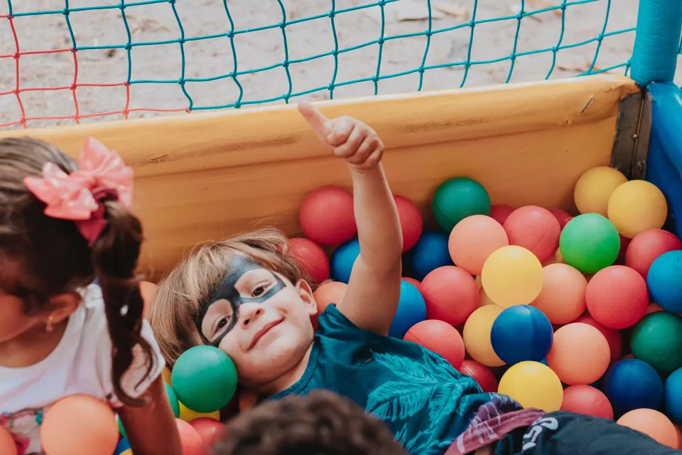 A child in face paint lying in a ball pit, giving a thumbs up
