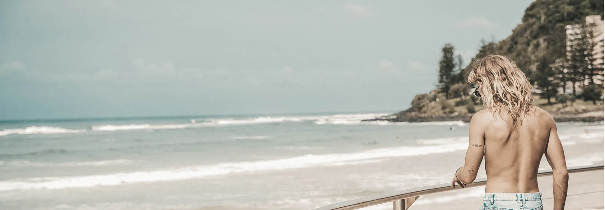 A long haired man seen from behind, leaning on a beachfront railing and looking along the surf towards a green headland