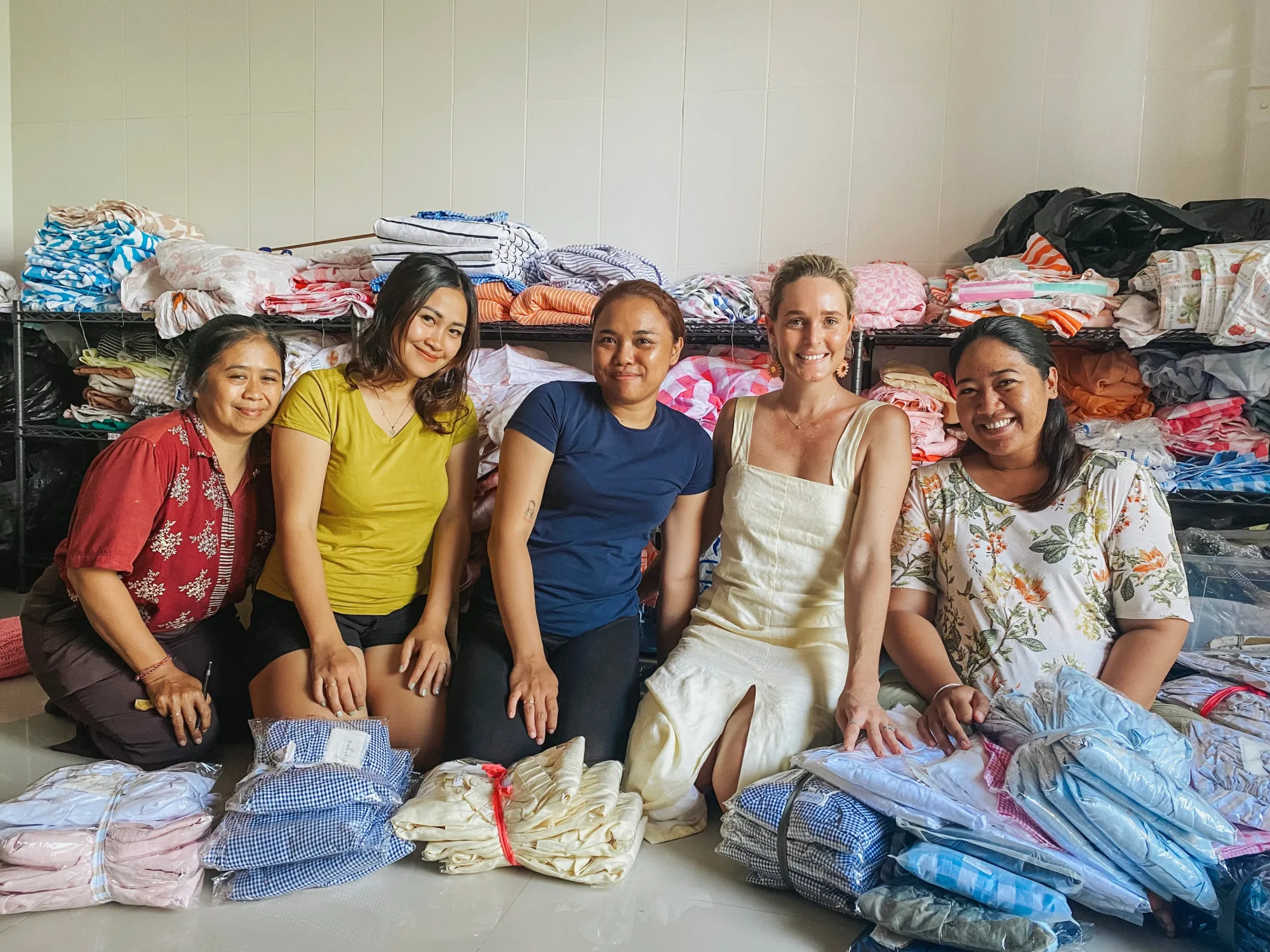 Five women kneeling on the floor among stacks of folded and packed linen garments, with shelves of fabric behind them
