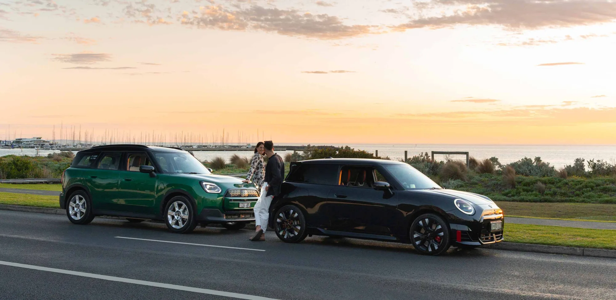 A green MINI and a black MINI parked on a waterfront road at sunset, with a couple standing between them and a marina behind