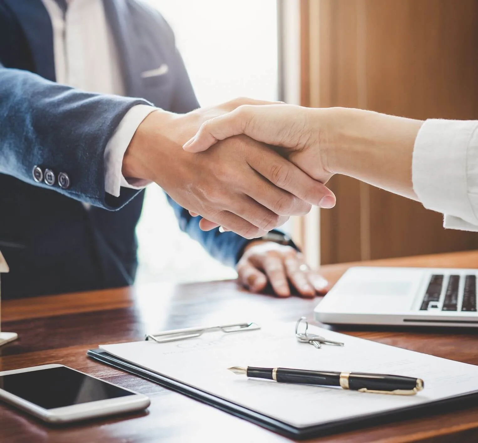 Two people shaking hands across a desk, with a signed document, a pen, a set of keys and a laptop in front of them
