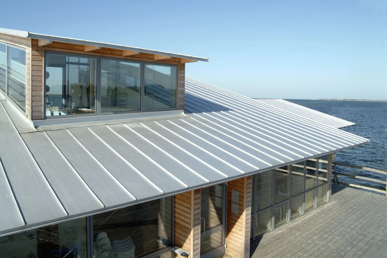 A standing seam metal roof on a timber and glass building beside open water under a clear blue sky
