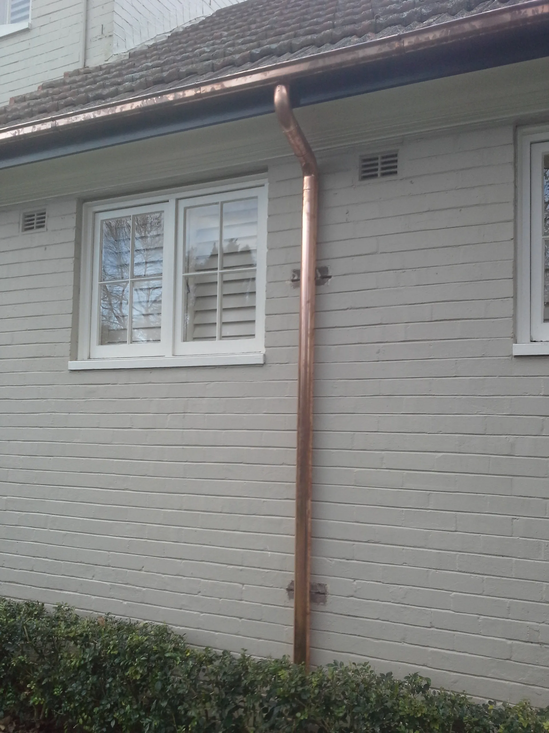 A copper downpipe running down a painted brick house wall beside a white window