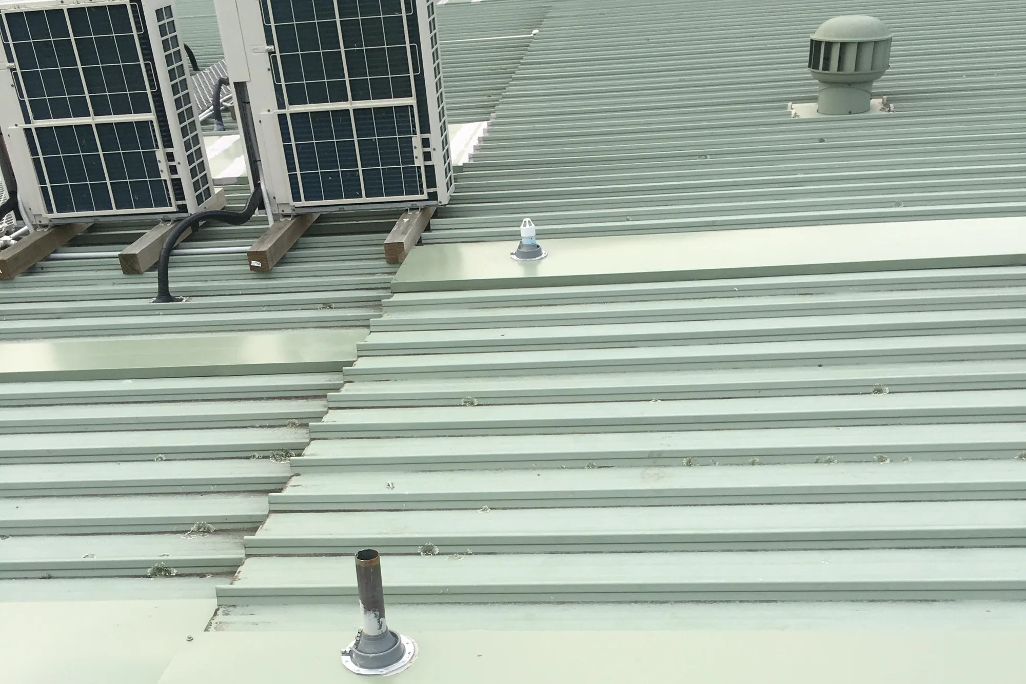A pale green metal roof with new sheeting, pipe flashings and a roof vent, air conditioning units standing on timber at one side
