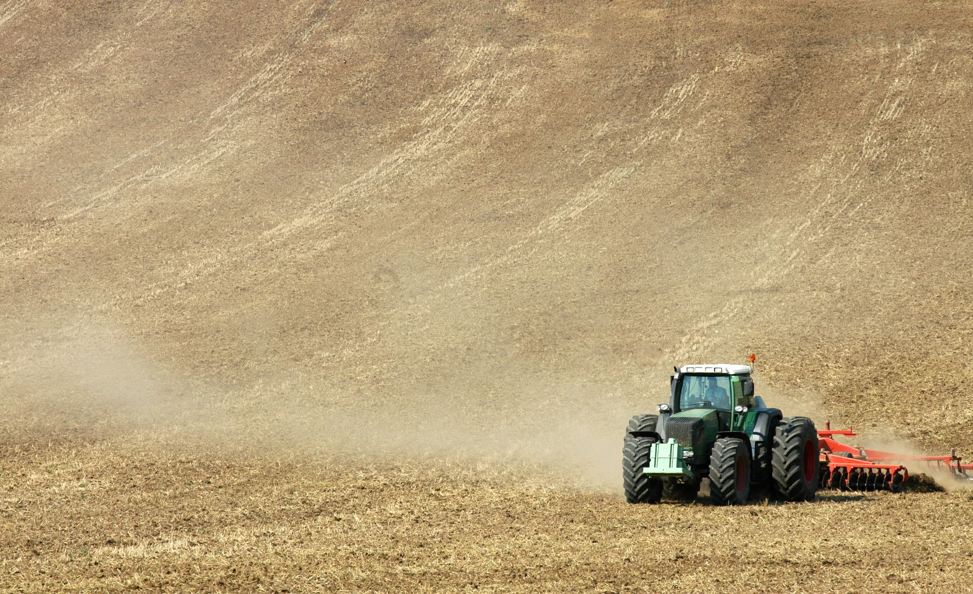 A green tractor pulling a disc harrow across a dry, dusty paddock