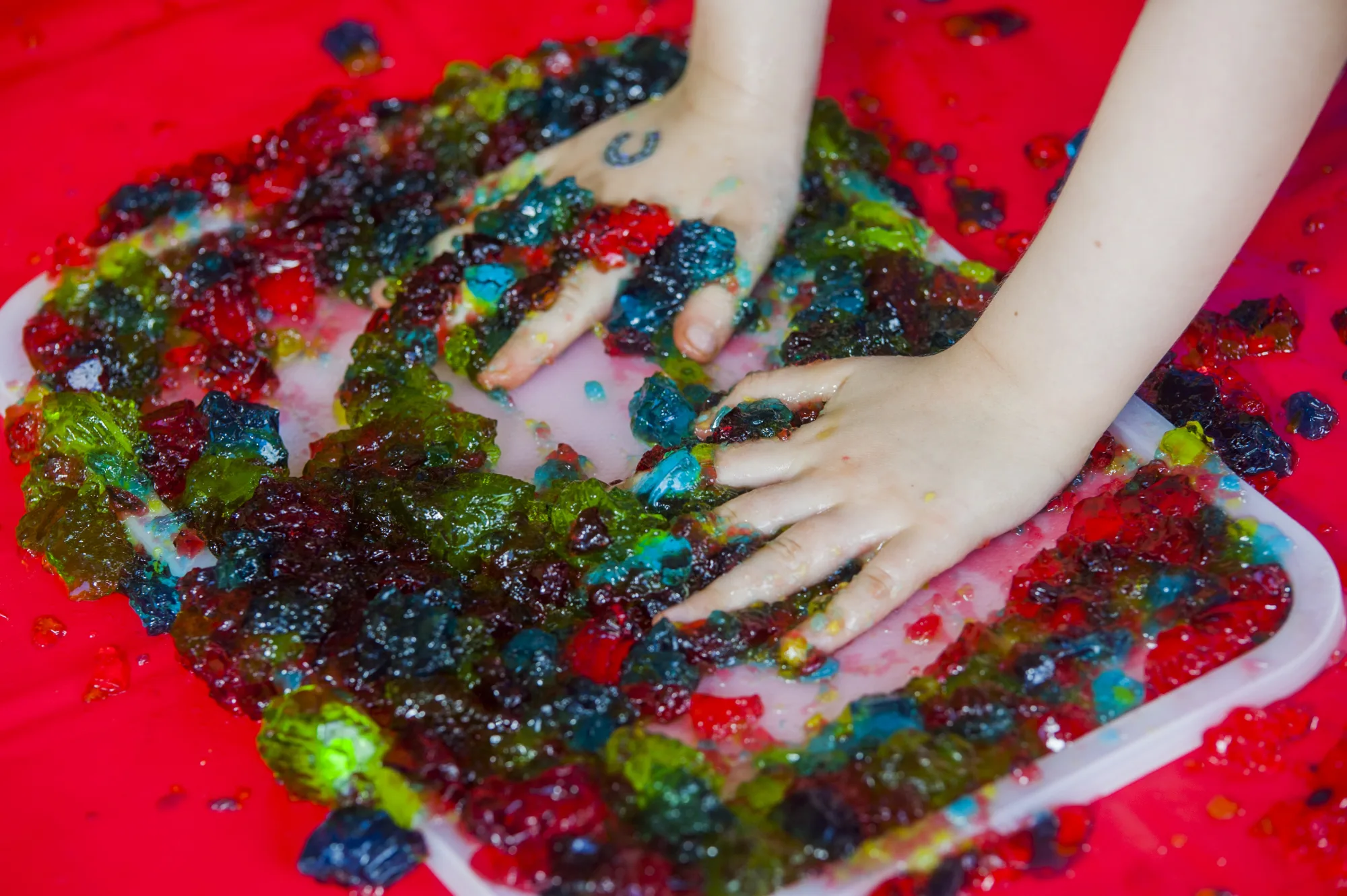 A child's hands pressing into bright coloured jelly in a tray on a red play mat