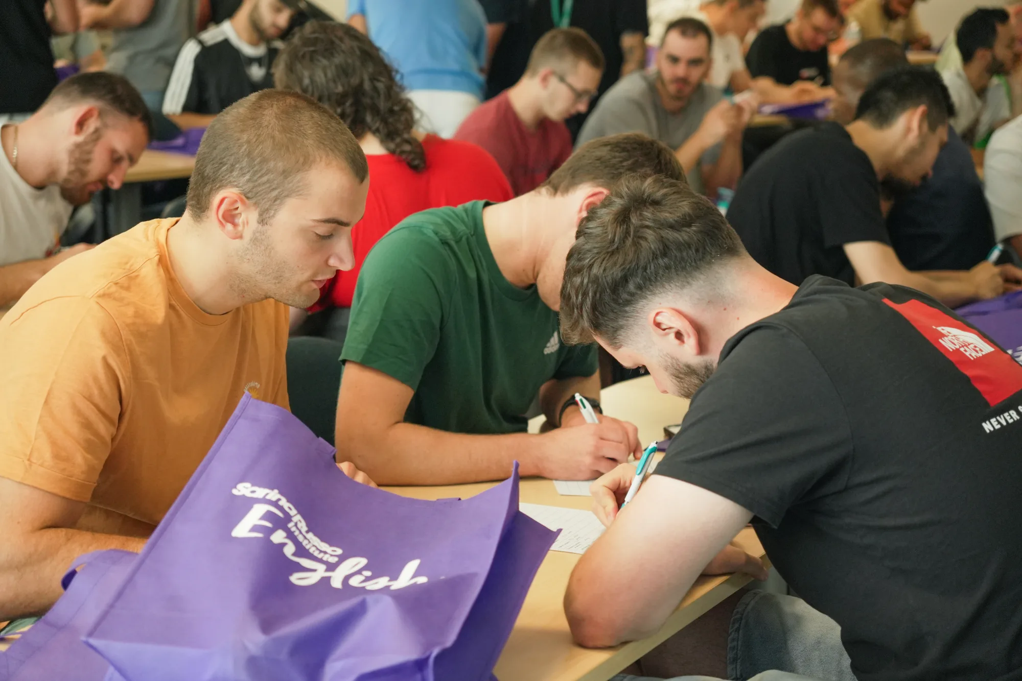 Students writing at a shared desk in a classroom, a purple Sarina Russo bag in front of them