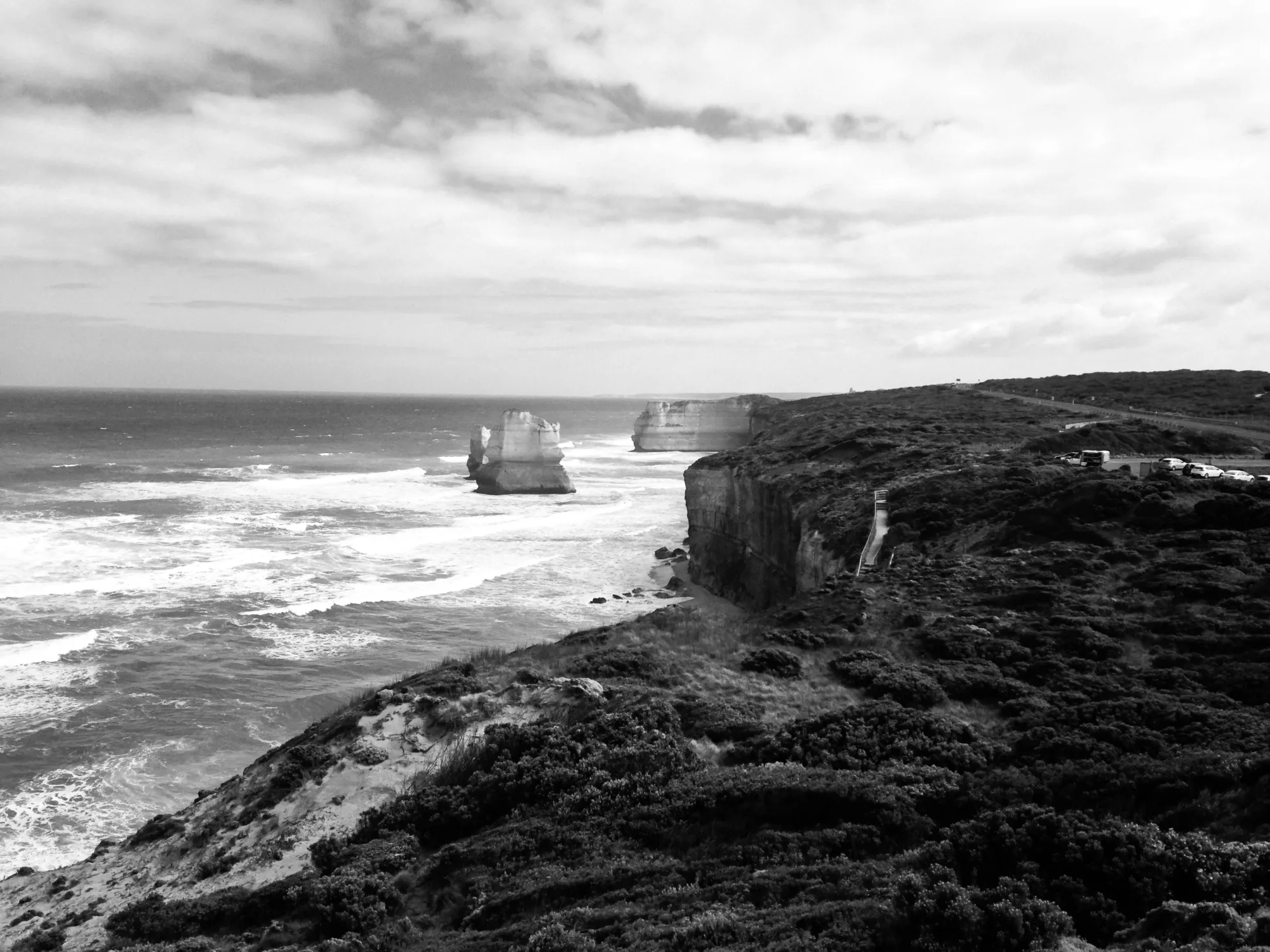 A black and white view along a clifftop coastline, limestone stacks standing in the surf below