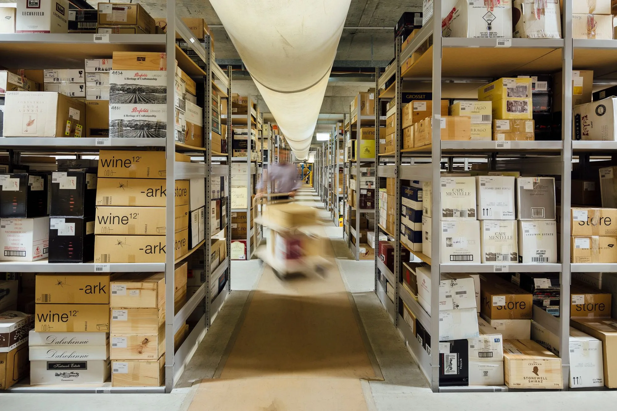 An aisle of Wine Ark storage shelving stacked with wine cases, a staff member wheeling a trolley down the middle